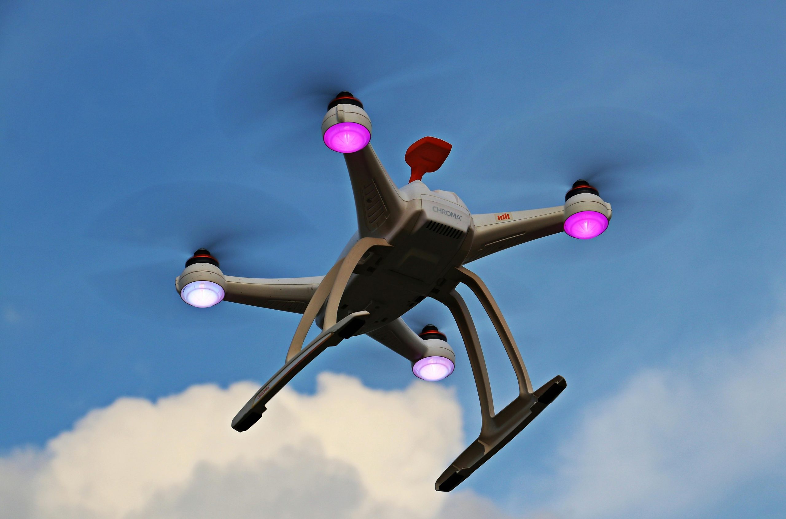 A quadcopter drone with propellers and lights flying high against a blue sky with clouds.